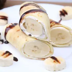 Rolowane naleśniki z bananem i bitą śmietaną, polane czekoladą. Obok ułożone plasterki banana z polewą.