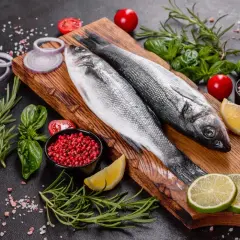 Dwie świeże ryby bass na desce, otoczone cytryną, limonką, pomidorkami, cebulą, rozmarynem i pieprzem.