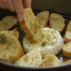 Ciepły serek camembert w okrągłej formie, otoczony kromkami pieczywa z ziołami. Ktoś zanurza kawałek chleba w rozpływającym się serze.