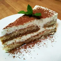 Pyszny kawałek najprostszego tiramisu, posypany kakao i udekorowany listkiem mięty, na białym talerzu.