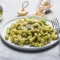 Pyszne pesto z makaronem fusilli, posypane parmezanem. Obok miseczka sosu, kawałki sera i orzeszki piniowe.
