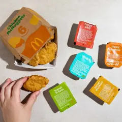 Ręka sięga po McNuggetsy z pudełka McDonald's, obok sosy: Tangy BBQ, Spicy Buffalo, Ranch, Sweet 'N Sour i Honey Mustard.