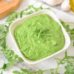 Aksamitne, zielone pesto z rukoli w białej miseczce, otoczone świeżymi listkami rukoli, czosnkiem i oliwą.