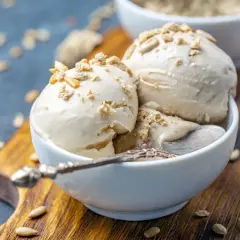 Trzy gałki lodów z mascarpone, posypane kruszonką i nasionami słonecznika, w białej miseczce na drewnianej desce.