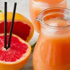 Orzeźwiający sok z grejpfruta w dzbanku i szklance, obok przekrojonych owoców z czarnymi słomkami.