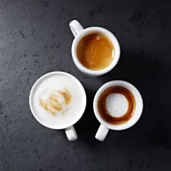 Trzy białe filiżanki z kawą: espresso, latte z wzorem i kawa macchiato na ciemnym tle.
