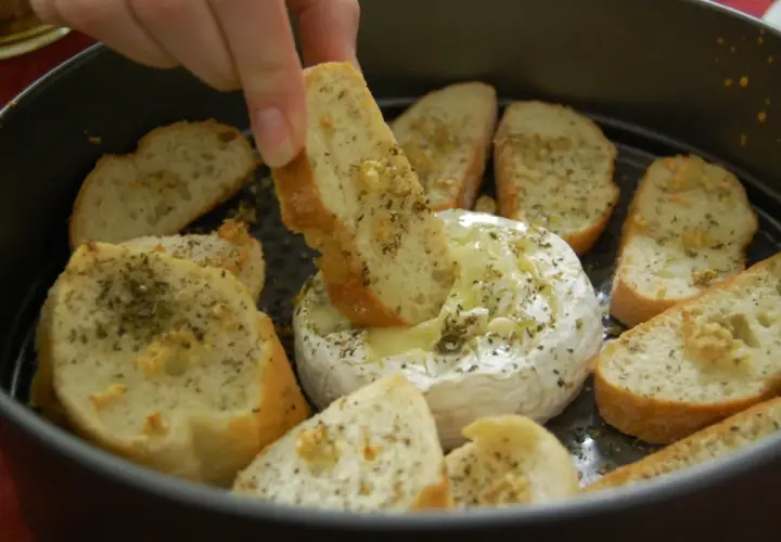Ciepły serek camembert w okrągłej formie, otoczony kromkami pieczywa z ziołami. Ktoś zanurza kawałek chleba w rozpływającym się serze.