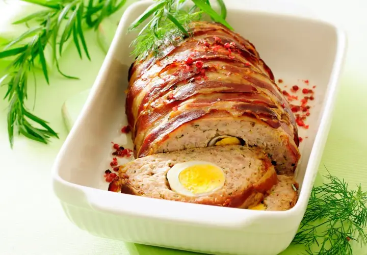 Najlepsza pieczeń rzymska, zawinięta w boczek, z jajkiem w środku, podana w białej brytfannie, ozdobiona ziołami i pieprzem.