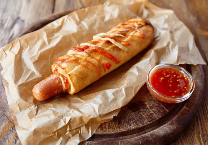 Pyszny hot dog francuski w bułce, polany keczupem i posypany serem, z sosem na boku.