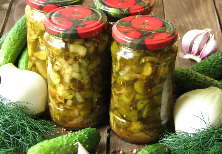 Słoiki z ogórkami w curry ze skórką, otoczone świeżymi ogórkami, cebulą, czosnkiem i koprem.