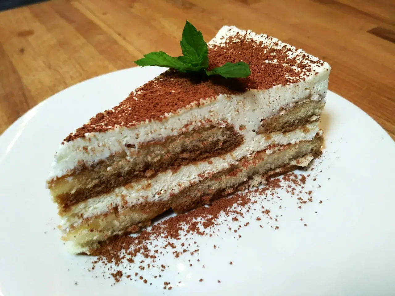 Pyszny kawałek najprostszego tiramisu, posypany kakao i udekorowany listkiem mięty, na białym talerzu.