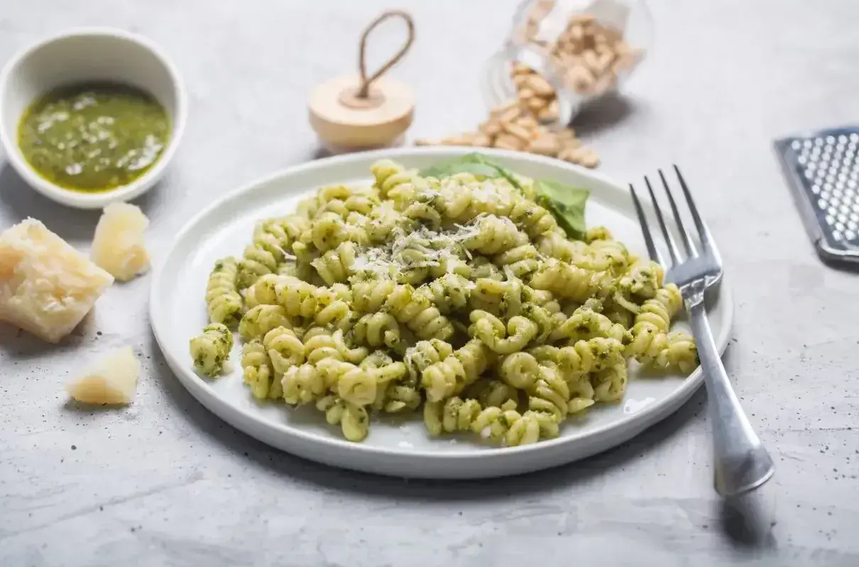 Pyszne pesto z makaronem fusilli, posypane parmezanem. Obok miseczka sosu, kawałki sera i orzeszki piniowe.