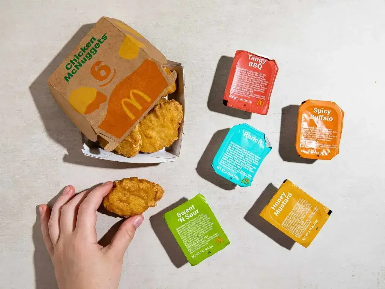 Ręka sięga po McNuggetsy z pudełka McDonald's, obok sosy: Tangy BBQ, Spicy Buffalo, Ranch, Sweet 'N Sour i Honey Mustard.