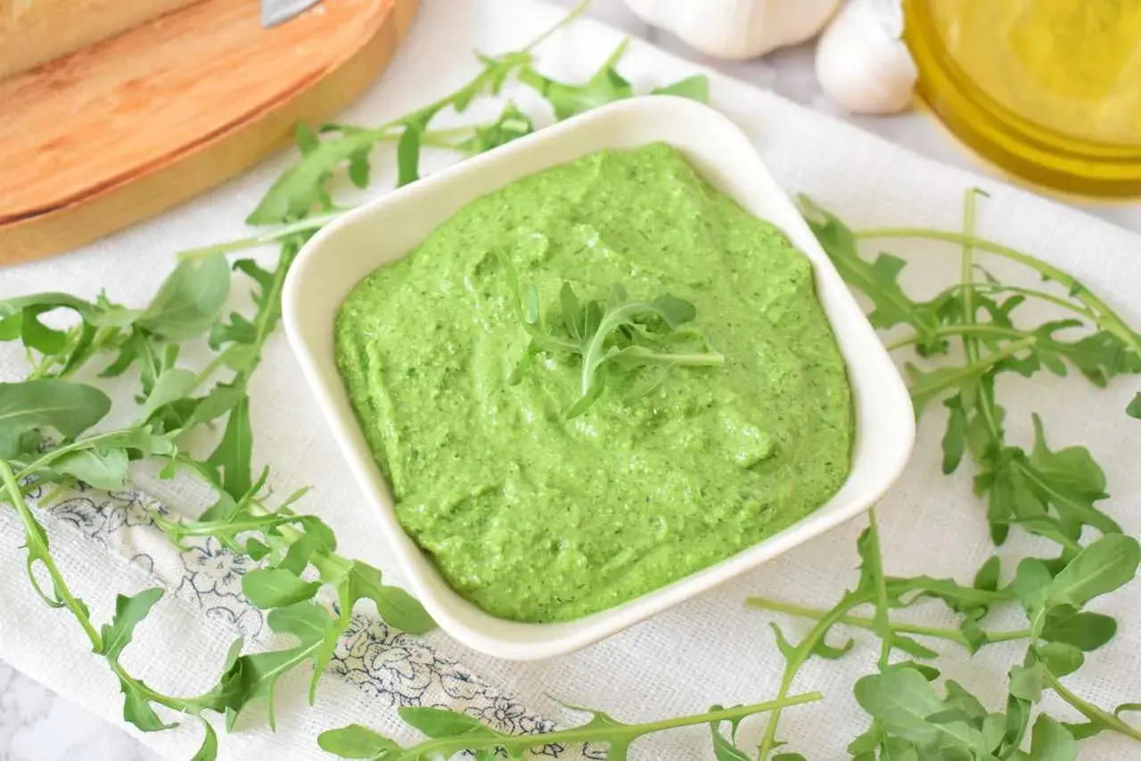 Aksamitne, zielone pesto z rukoli w białej miseczce, otoczone świeżymi listkami rukoli, czosnkiem i oliwą.