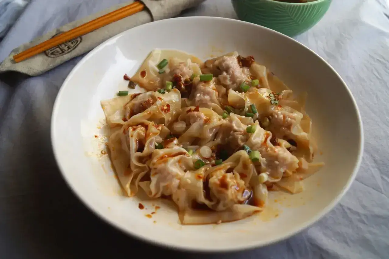 Pyszne ciasto wonton w pikantnym sosie, posypane szczypiorkiem i sezamem. Obok pałeczki i miseczka z sosem.