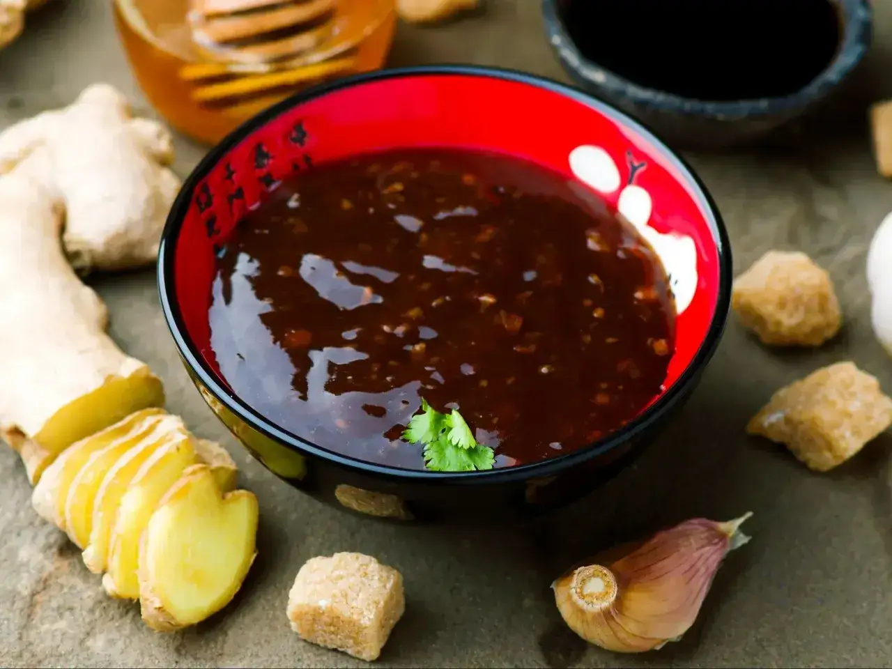 Sos teriyaki przepis: miska pełna aromatycznego sosu, obok świeży imbir, czosnek, brązowy cukier i miód. Idealne składniki do domowego sosu teriyaki.