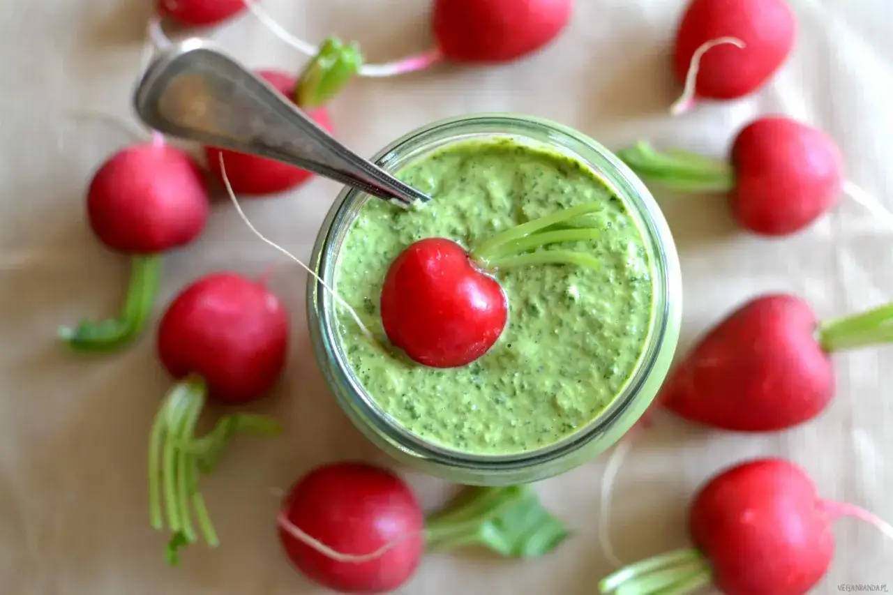 Pesto z liści rzodkiewki w słoiku, ozdobione rzodkiewką i listkami. Wokół rozłożone świeże rzodkiewki.